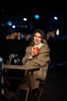 lonely beautiful girl in the evening at a table in a street cafe with a glass of coffee in her hands, night, bokeh photo