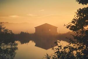 Old barn on lake shore on misty sunrise photo