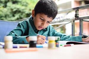 Smart Indian little boy perform thumb painting with different colourful water colour kit during the summer vacations, Cute Indian Kid doing colourful thumb painting drawing on wooden table photo