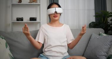 Selective focus, Relaxed young woman wear VR goggles meditating doing a lotus pose at home sitting on sofa and experiencing virtual reality in living room photo