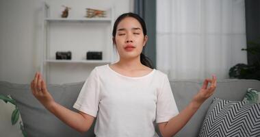 selectivo enfocar, contento joven mujer meditando haciendo un loto actitud a hogar con ojos cerrado, relajante cuerpo y mente solo en sofá en el vivo habitación, relajación estilo de vida foto