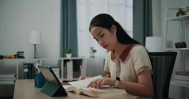 selectivo enfocar, lado vista, hembra inalámbrico auriculares escritor sentado a escritorio escritura información en diario cuaderno mientras leyendo libro a hogar, creativo pensamientos a diario, idea y inspiración foto