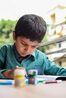 Smart Indian little boy perform thumb painting with different colourful water colour kit during the summer vacations, Cute Indian Kid doing colourful thumb painting drawing on wooden table photo