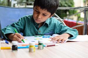 Smart Indian little boy perform thumb painting with different colourful water colour kit during the summer vacations, Cute Indian Kid doing colourful thumb painting drawing on wooden table photo