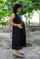 A pregnant Indian lady poses for outdoor pregnancy shoot and hands on belly, Indian pregnant woman puts her hand on her stomach with a maternity dress at society park, Pregnant outside maternity shoot photo