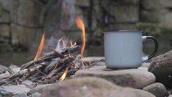 A mug on a stone near a fire on the river bank, close-up. Steam rises from the mug. Travel concept. 4k video