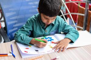 Smart Indian little boy perform thumb painting with different colourful water colour kit during the summer vacations, Cute Indian Kid doing colourful thumb painting drawing on wooden table photo