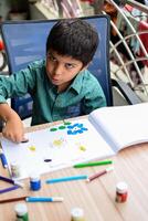 Smart Indian little boy perform thumb painting with different colourful water colour kit during the summer vacations, Cute Indian Kid doing colourful thumb painting drawing on wooden table photo