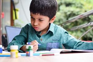 Smart Indian little boy perform thumb painting with different colourful water colour kit during the summer vacations, Cute Indian Kid doing colourful thumb painting drawing on wooden table photo