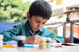 Smart Indian little boy perform thumb painting with different colourful water colour kit during the summer vacations, Cute Indian Kid doing colourful thumb painting drawing on wooden table photo