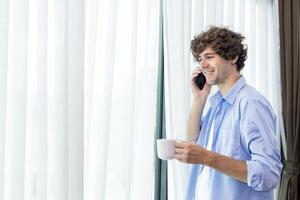 hermoso caucásico persona de libre dedicación utilizando móvil teléfono y Bebiendo café taza mirando fuera el ventana a su hogar Departamento en el Mañana antes de yendo a trabajo para vida equilibrar con Copiar espacio foto