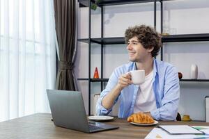 caucásico hombre es trabajando a su hogar oficina mientras Bebiendo café y comiendo cuerno para desayuno para Lanza libre y empresario negocio foto