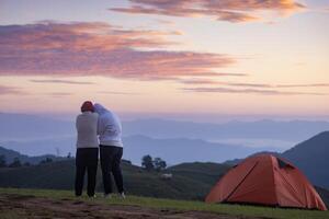 Couple is hugging each other by the tent during overnight camping while looking at the beautiful scenic sunset over the mountain for outdoor adventure vacation travel photo