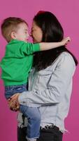 adorable pequeño chico abrazando su madre y jugando alrededor, teniendo divertido en contra rosado antecedentes. joven niñito posando con su mamá participación a él, riendo y siendo alegre. cámara b. video