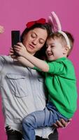 Lovely small child taking pictures with his mother on smartphone, trying to capture fun and cute moments against pink background. Little boy being playful and fooling around with phone. Camera B. video