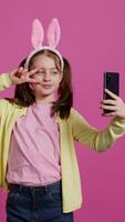 Confident happy young girl taking photos on smartphone webcam, feeling cute with her pigtails and bunny ears. Joyful smiling toddler takes pictures and fooling around in studio. Camera B. video