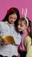 Mother reading fairytale story to her sweet clever daughter, enjoying fiction book with novels against pink background. Cheerful curious schoolgirl with bunny ears listening to her mom. Camera B. video