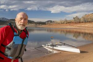 ambiental retrato de un mayor hombre vistiendo traje de buceo y vida chaqueta con un remo cáscara en lago apuntalar foto