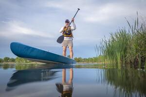 mayor masculino palista es remar un estar arriba paddleboard en un calma lago en primavera, rana perspectiva desde un acción cámara a agua nivel foto