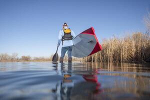 senior male paddler is launching a stand up paddleboard on a calm lake in early spring, frog perspective from an action camera at water level photo