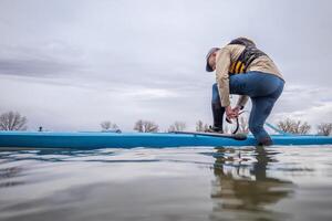 mayor masculino es adjuntando un la seguridad Correa antes de remar un estar arriba paddleboard en un lago en temprano primavera, rana perspectiva desde un acción cámara a agua nivel foto