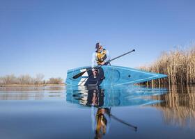 senior male paddler is launching a stand up paddleboard on a calm lake in early spring, frog perspective from an action camera at water level photo