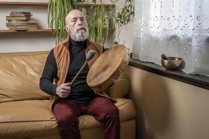 senior man is playing a native American style, shaman drum with a beater at home, lifestyle and hobby concept photo