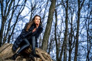 al aire libre retrato de joven bonito hermosa mujer en frío soleado día en parque. sensual morena posando en piel Saco sentado en un rock en parque foto