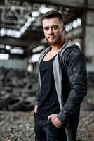 Brutal young handsome man in stylish clothes posing at abandoned broken warehouse background. Vertical shot photo