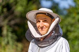 Head and shoulders portrait of a senior beekeeper in white protective hat with net. Blurred background of green trees. Closeup. photo