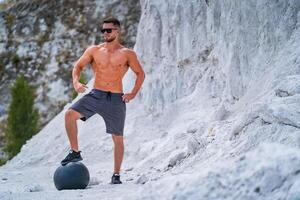 Brutal strong bodybuilder posing outdoor. Photoshoot in a quarry. Outdoor sports concept. Front view. Posing with leg on fitball. White landscape photo