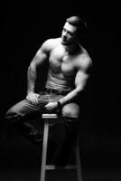 Muscular and fit young bodybuilder fitness male model posing on chair. Black and white photo. Full size portrait. photo