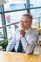 Mature businessman looking in front. Thinking over ideas. Sitting near the window in cafe. Panoramic city view background. Business photo. photo