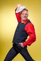 Old smiling grey-haired man in eyeglasses holding fan of dollars over head in dancing pose. Human emotions and facial expressions photo