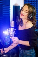 Focused pretty singer with headphones in front of the microphone recording a song in studio. Portrait of a beautiful young woman singing into the mic with headphones. Close-up photo