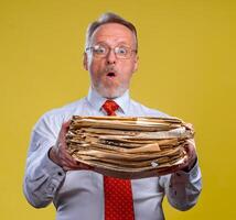 Frustrated executive worker over yellow background overworked holding piles of documents paper work. photo