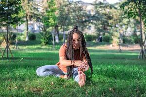 hermosa joven mujer con largo africano trenzas es haciendo yoga fuera de en un parque. concepto de sano estilo de vida. foto
