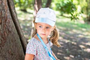 pequeño niña en un floral vestir y un béisbol gorra en pie cerca un árbol en el parque foto