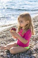 joven niña con largo rubio pelo sentado en un playa con mar ver y comiendo un rojo manzana. foto
