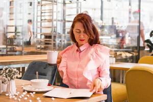 encantador joven mujer en rosado camisa disfrutando comiendo desayuno a cafetería. foto