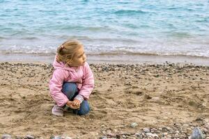 Happy little girl playing with colorful pebbles on sandy beach by ocean waves on sunny summer day photo