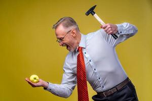 Strong business man wearing shirt holding hammer pointing at apple isolated on yellow background. Side view. photo