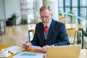 Mature businessman sitting at table with laptop reading printed documents. Sitting near the window in office. Panoramic city view background. Business photo. photo