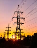 High voltage pylons towering over the trees and bushes. Dark tower silhouettes facing light pinky clouds in the orange sky. photo