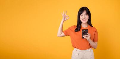 Energetic young Asian woman in her 30s, wearing orange shirt, using smartphone with okay hand sign on vibrant yellow background. New mobile app concept. photo