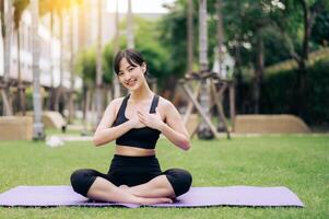 Portrait fit pretty young asian woman 30s wearing black sportswear holding hands on chest while listening relax music and sitting on yoga mat in public park. Healthcare wellness concept. photo