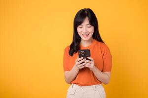 Smiling Asian woman in her 30s using smartphone, dressed in orange shirt, on vibrant yellow background. Mobile tech concept. photo