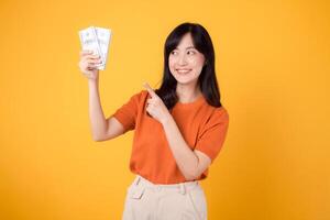 Energetic Asian woman in her 30s, pointing finger to cash money dollars, standing on vibrant yellow backdrop. photo