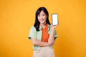 nuevo móvil solicitud con joven asiático mujer 30, vestido en naranja camisa y verde saltador, presentación teléfono inteligente pantalla en amarillo estudio antecedentes. nuevo móvil dispositivo y en línea compras concepto. foto