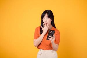 intrigado asiático mujer en su 30, vistiendo naranja camisa, utilizando teléfono inteligente con puño arriba mano firmar en vibrante amarillo antecedentes. fascinante nuevo aplicación exploración. foto